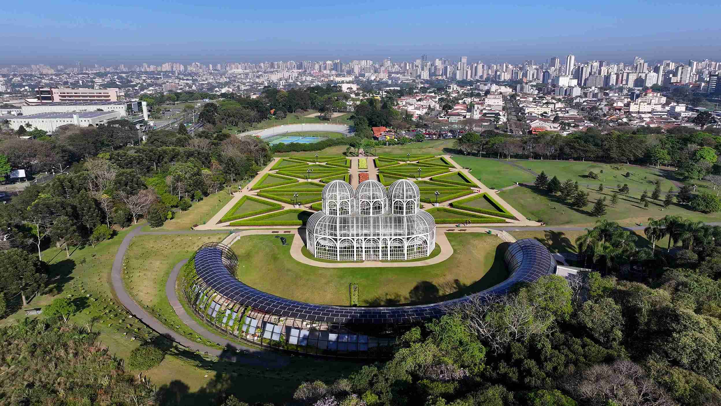 Vista aérea do Jardim Botânico de Curitiba, um dos pontos turísticos mais icônicos da cidade. A imagem destaca a harmoniosa integração entre áreas verdes e a malha urbana ao fundo, representando o crescimento planejado e valorização do mercado imobiliário em Curitiba.